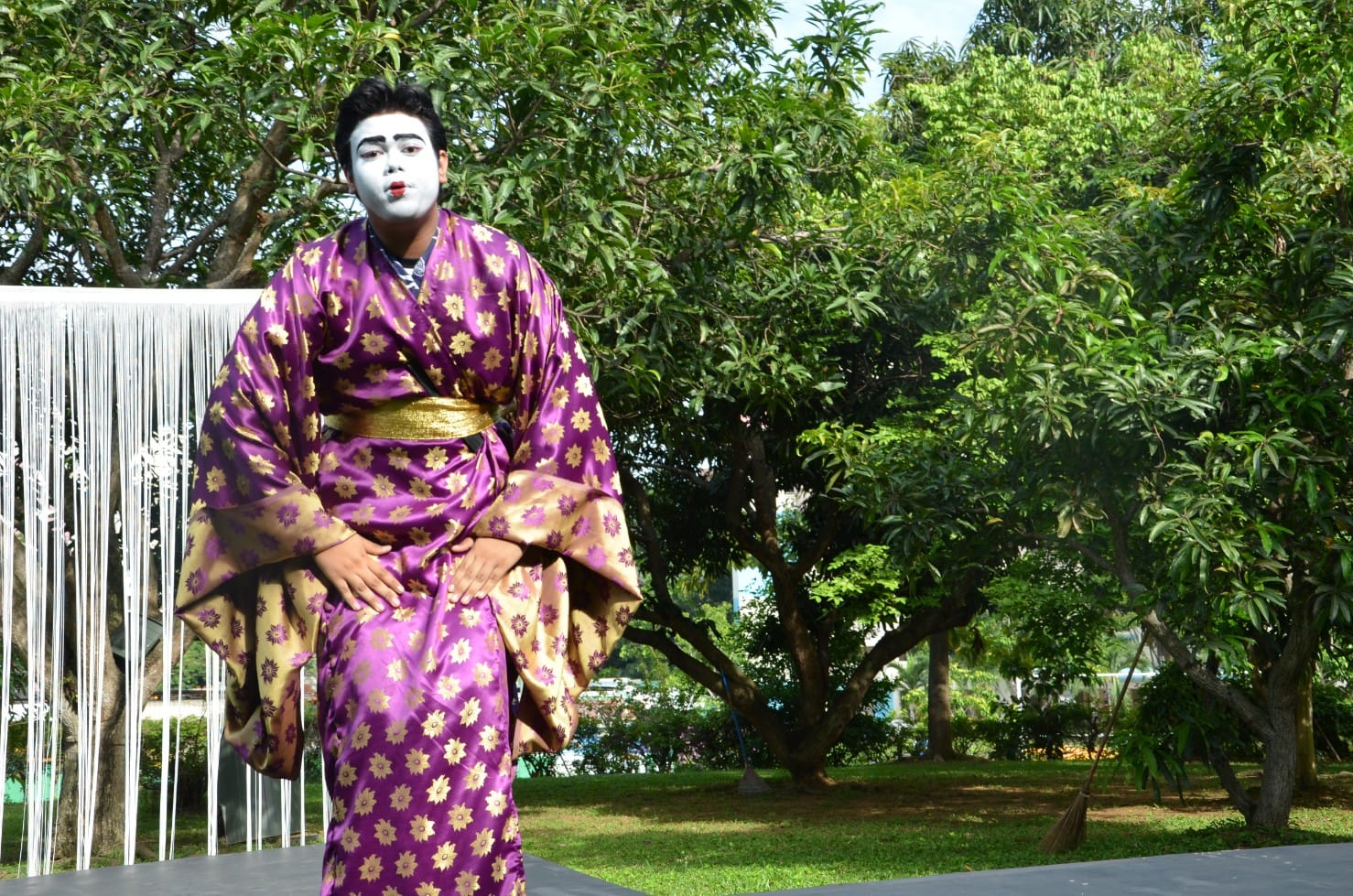 Performer in a purple and gold floral kimono with white face makeup stands outdoors among green trees.